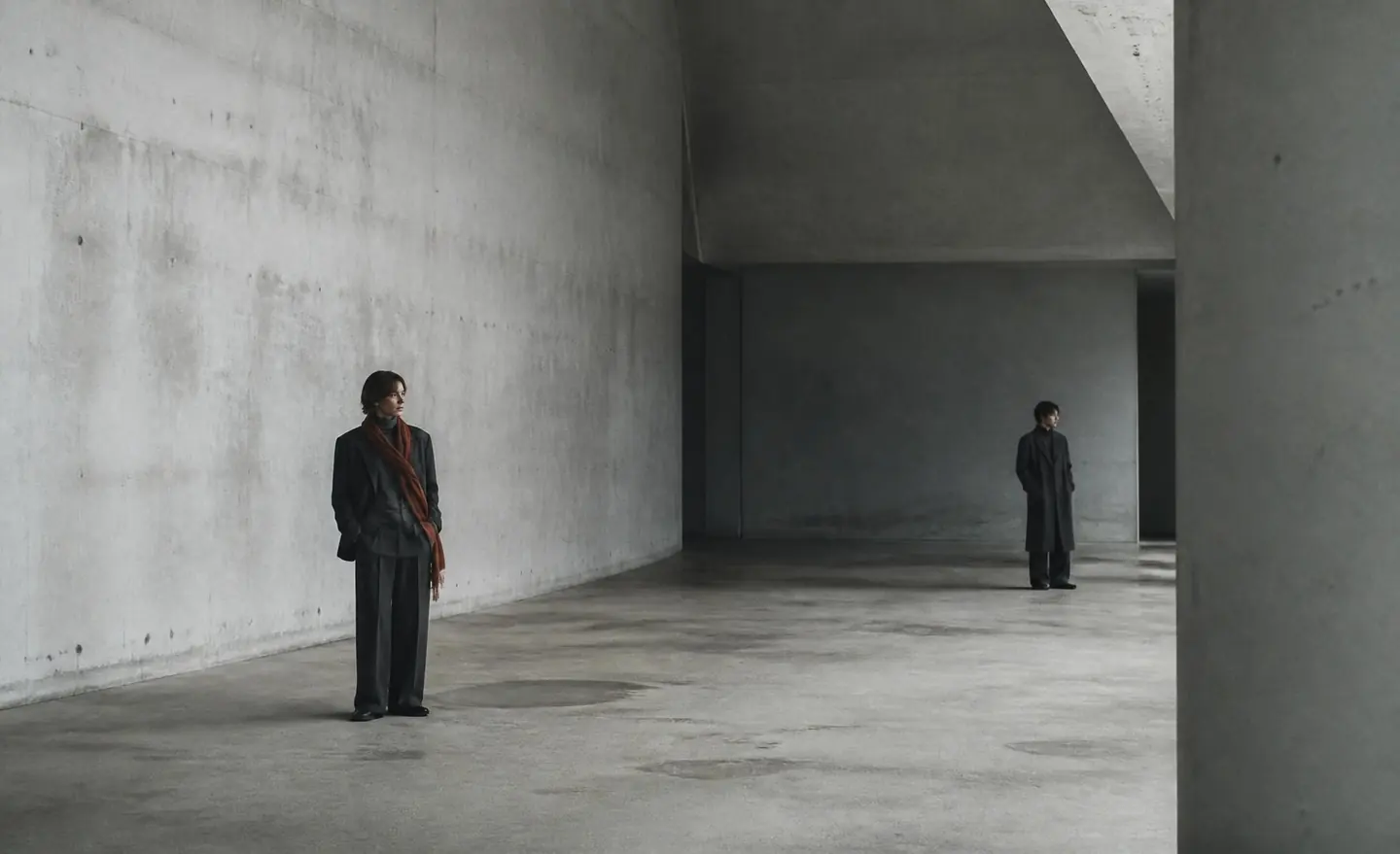 Two figures in charcoal wool coats standing far apart in a vast concrete hall, one wearing a taupe scarf.