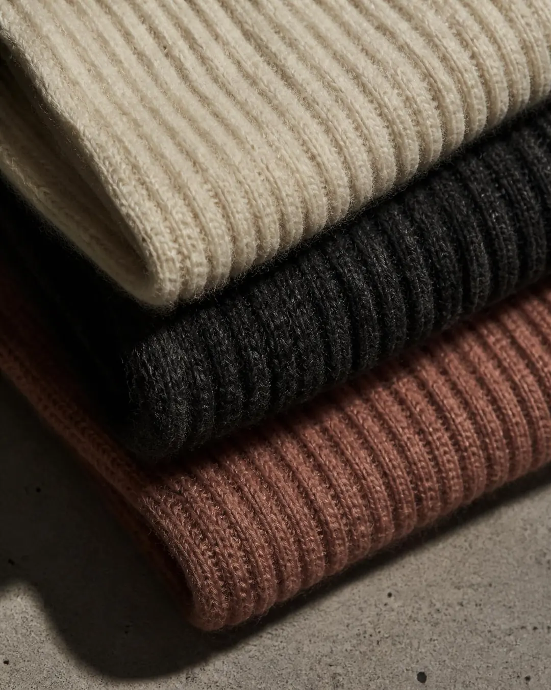 Close crop of stacked ribbed knit swatches in ecru, charcoal and taupe on a warm stone surface.