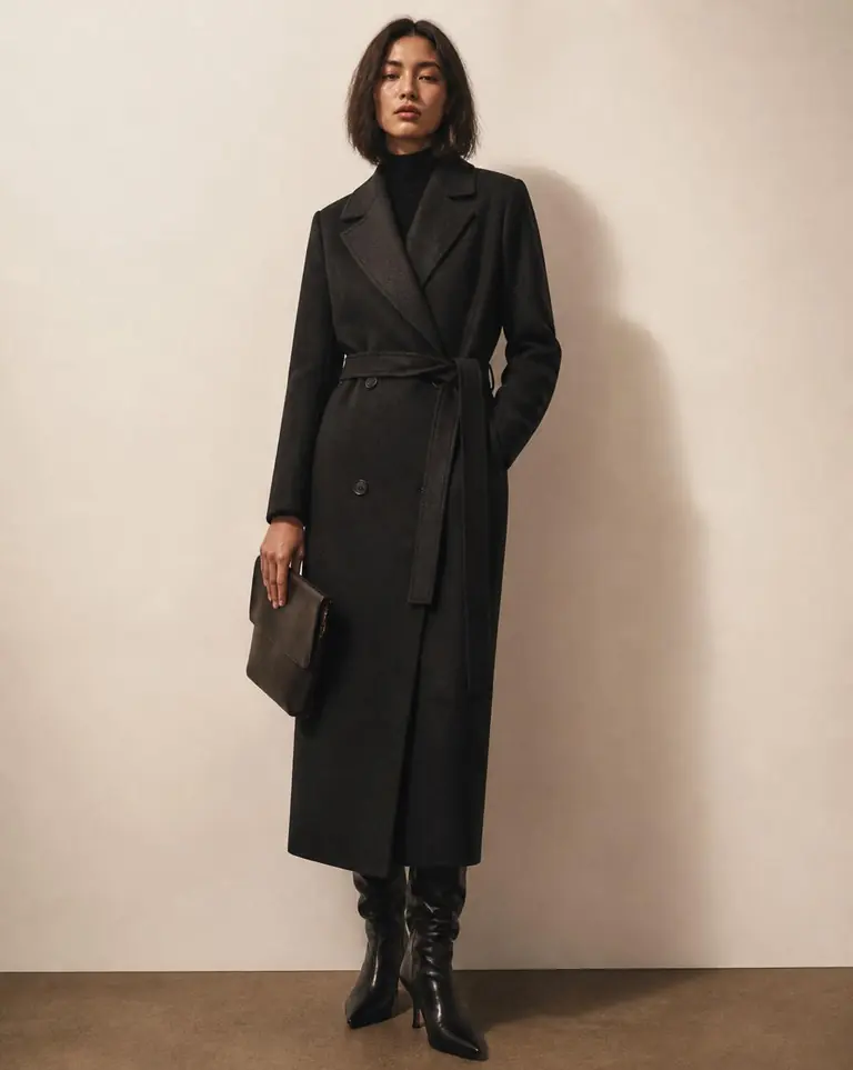 Model wearing a charcoal black tailored wool coat against a warm off-white wall.